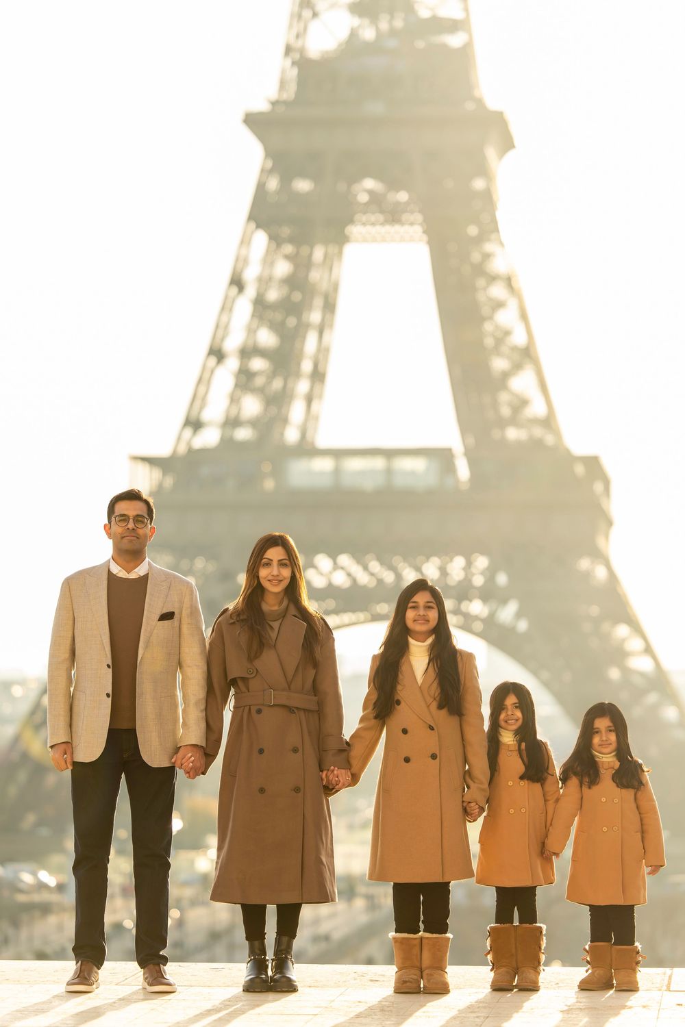 A family in coordinated brown and tan coats stands in line by height with the Eiffel Tower visible in the background.