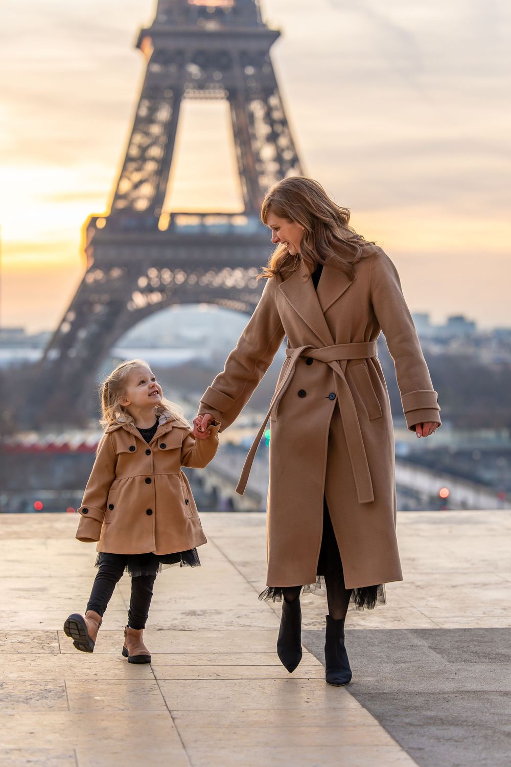 A parent and child wearing matching camel coats hold hands while walking near the illuminated Eiffel Tower at sunset.