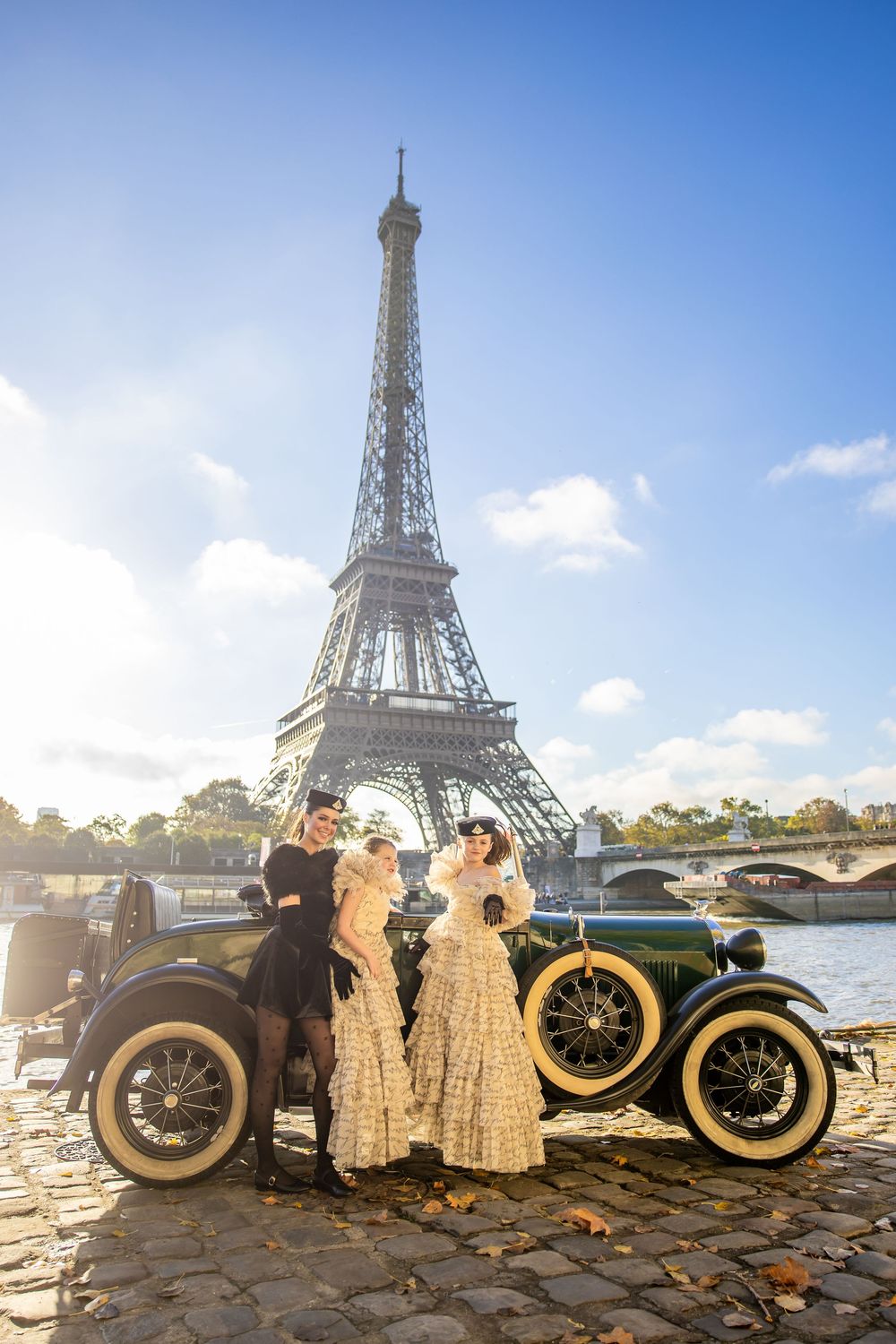Vintage car and people in period dress pose in front of the Eiffel Tower on a sunny day in Paris.