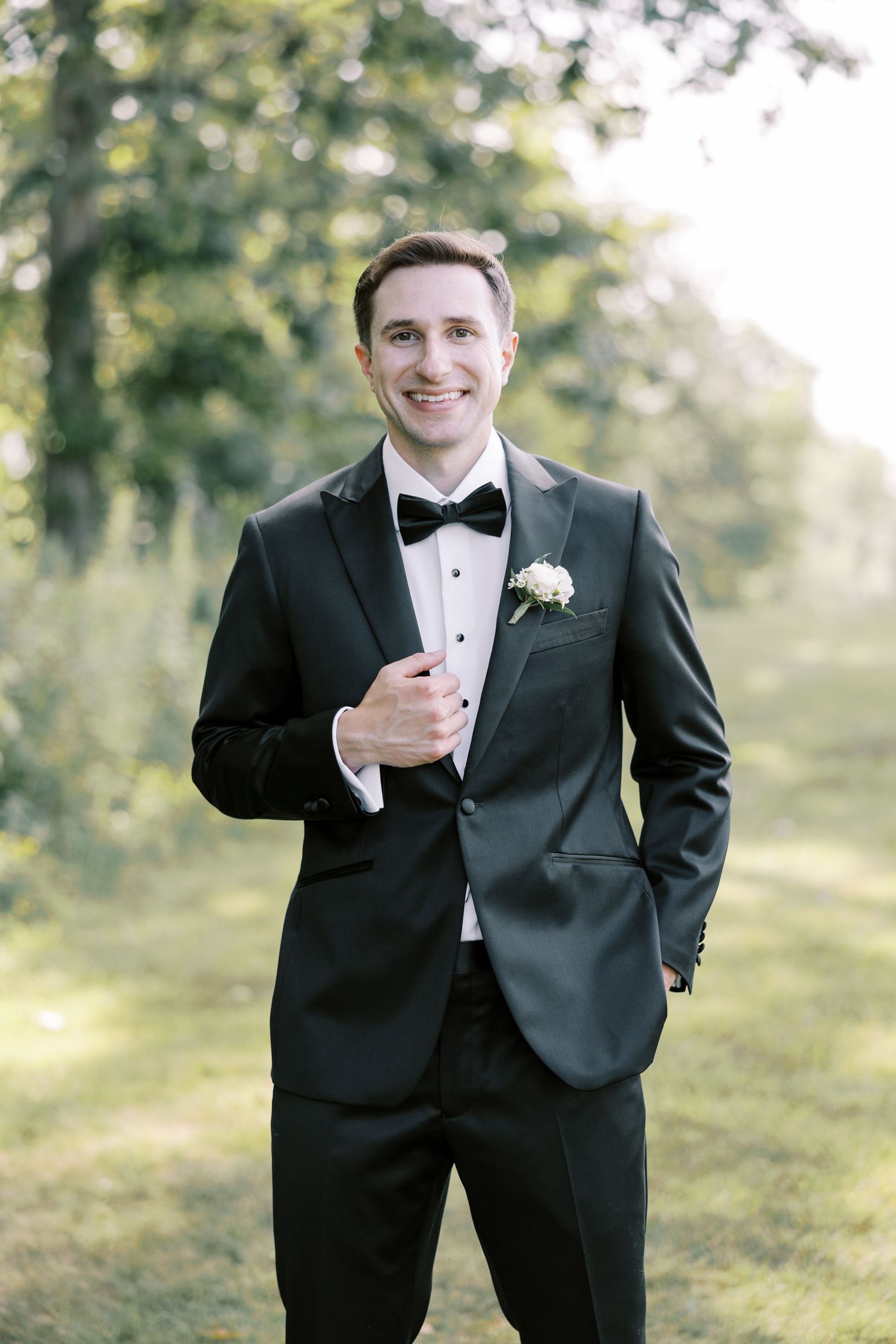 A groom in a classic black tuxedo and bowtie poses outdoors in a sunlit garden setting with a natural backdrop.