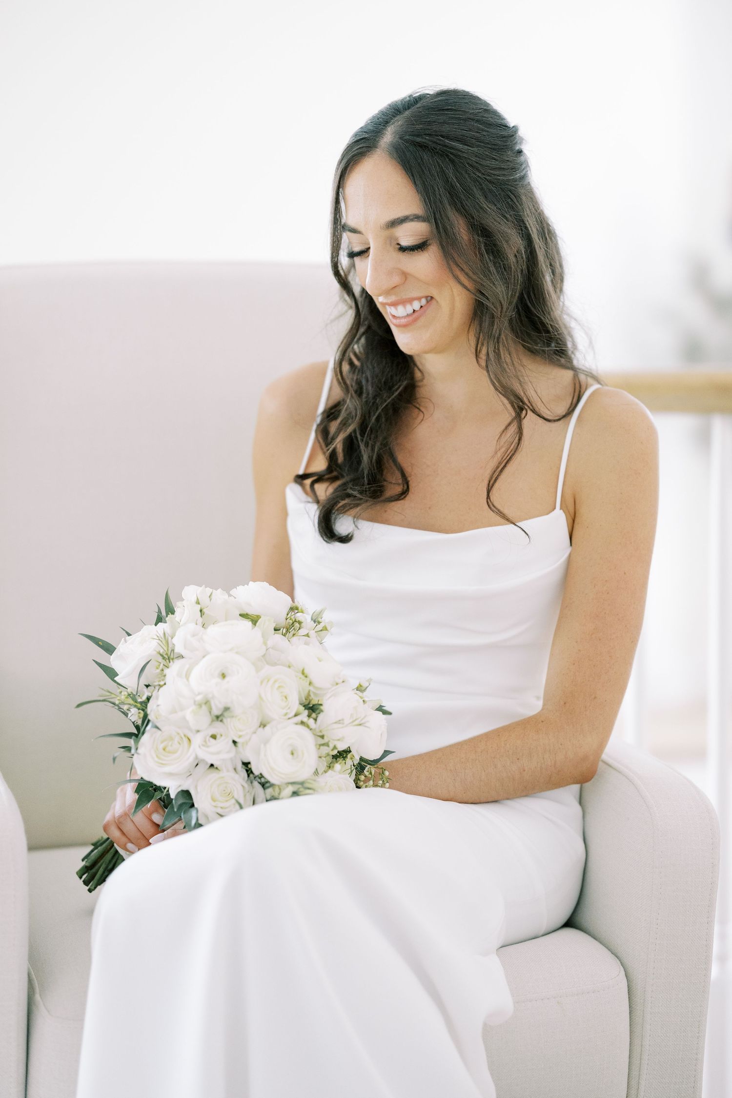 Elegant bride in white dress sits gracefully with white floral bouquet in natural light setting.