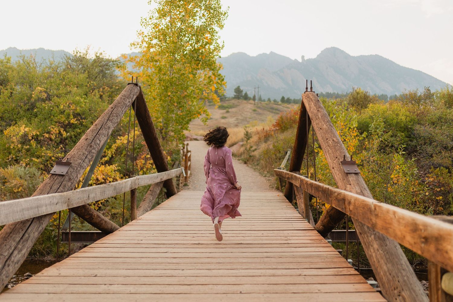 A wooden footbridge spans across mountains in autumn with someone in a flowing pink dress walking away.