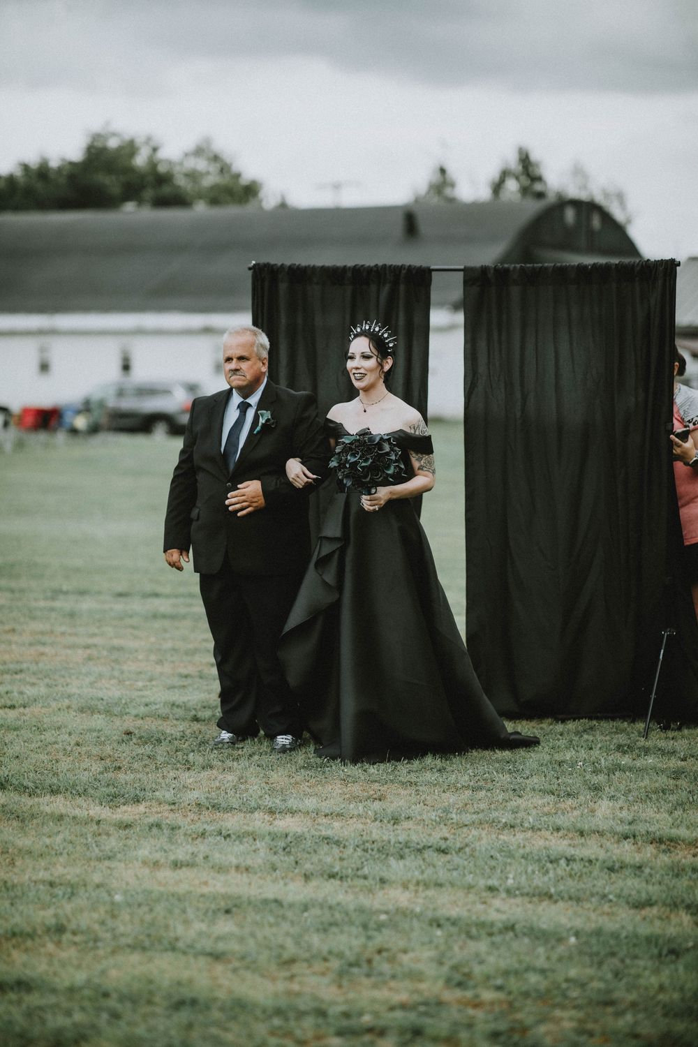 At an outdoor wedding ceremony, attendees walk across a grassy field next to a black curtain backdrop and barn.