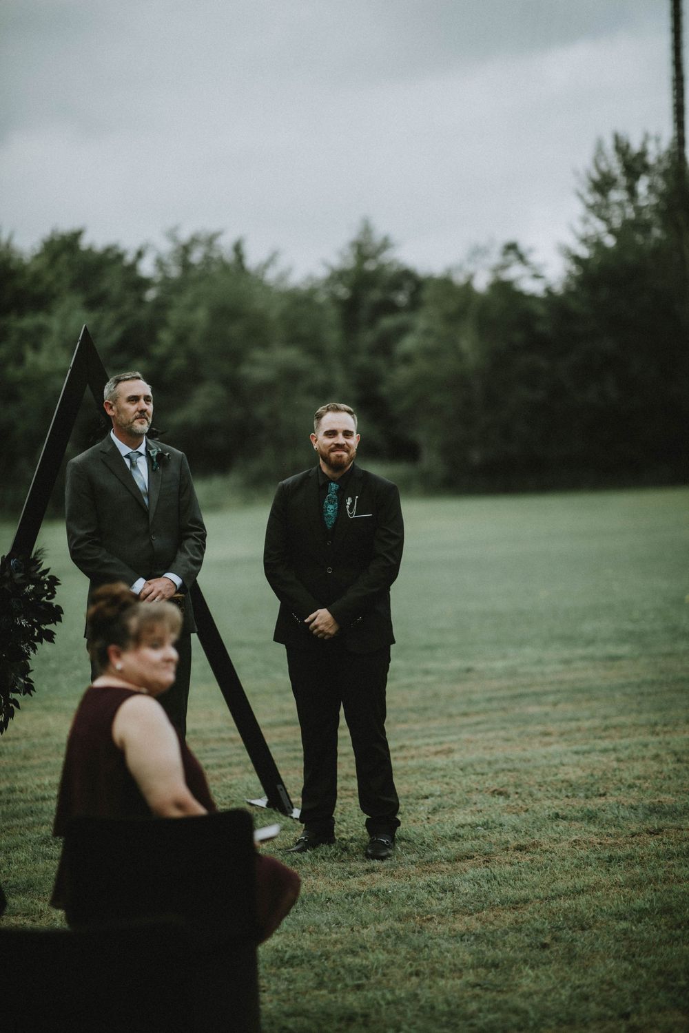 Two individuals in dark suits standing on a green lawn while someone in black sits nearby on an overcast day.