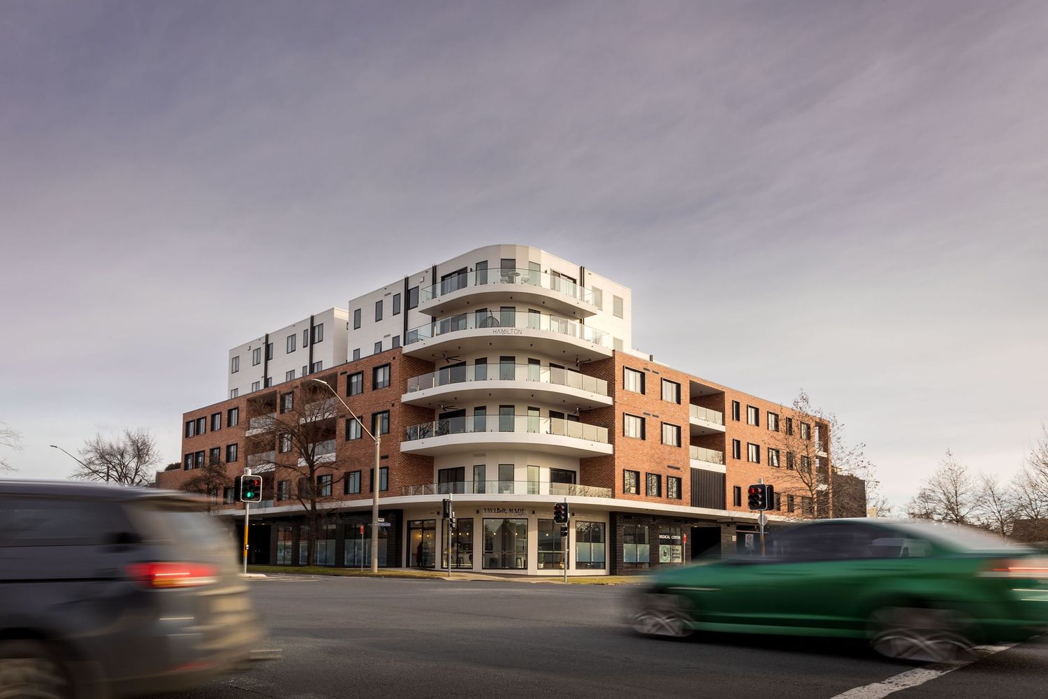 Modern apartment building with brick facade and white balconies captured with motion-blurred cars passing in the foreground.