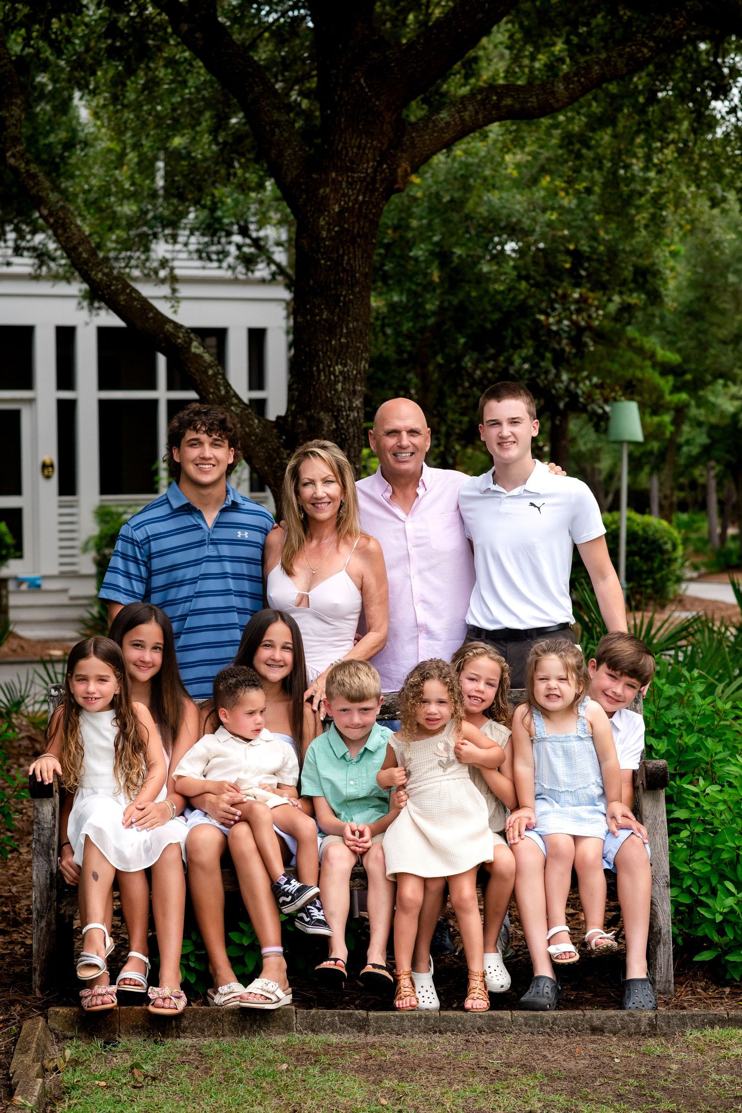 Large family group poses together outdoors while sitting on wooden bench under trees and greenery.