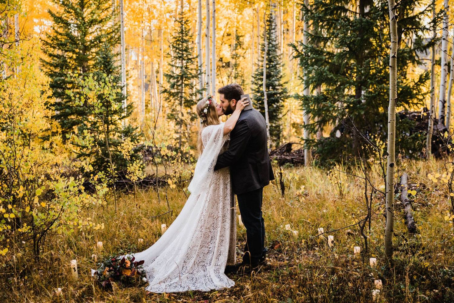 Romantic wedding couple embraces in a golden autumn forest setting with evergreen trees at sunset.