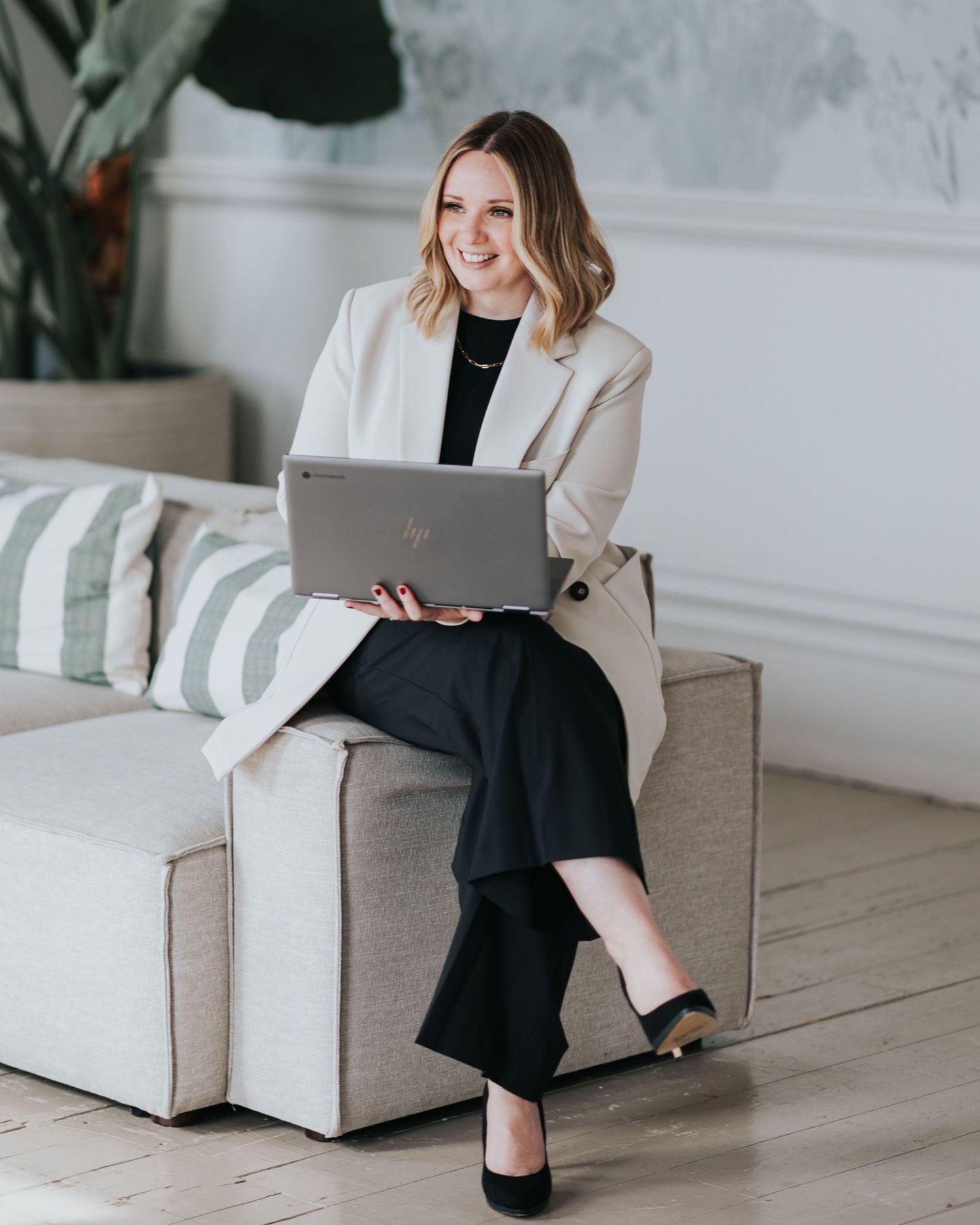 Professional woman in business attire sits on gray couch with laptop in bright modern office setting.