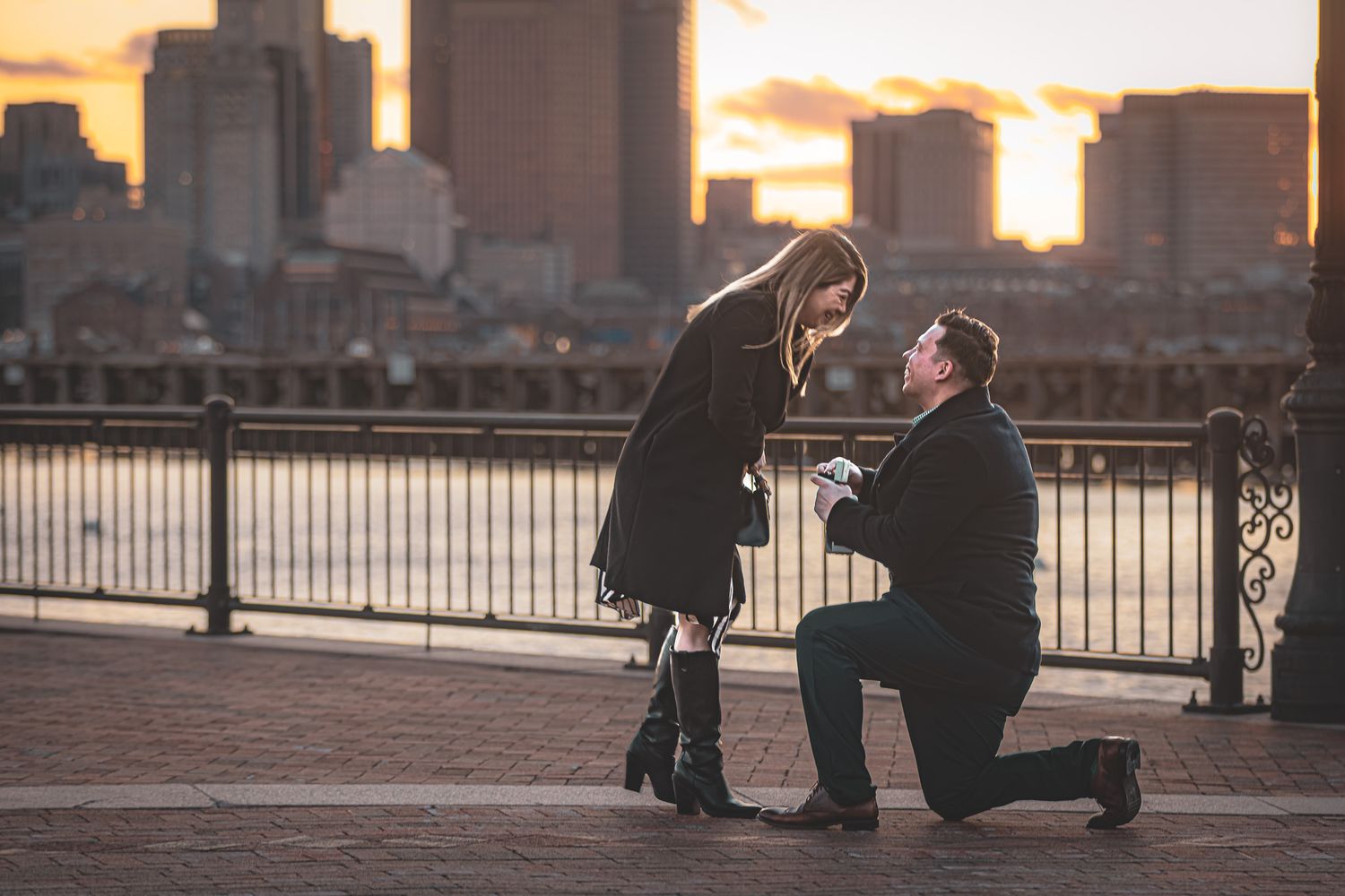 A silhouetted couple sharing a romantic proposal moment at sunset along a waterfront city backdrop.