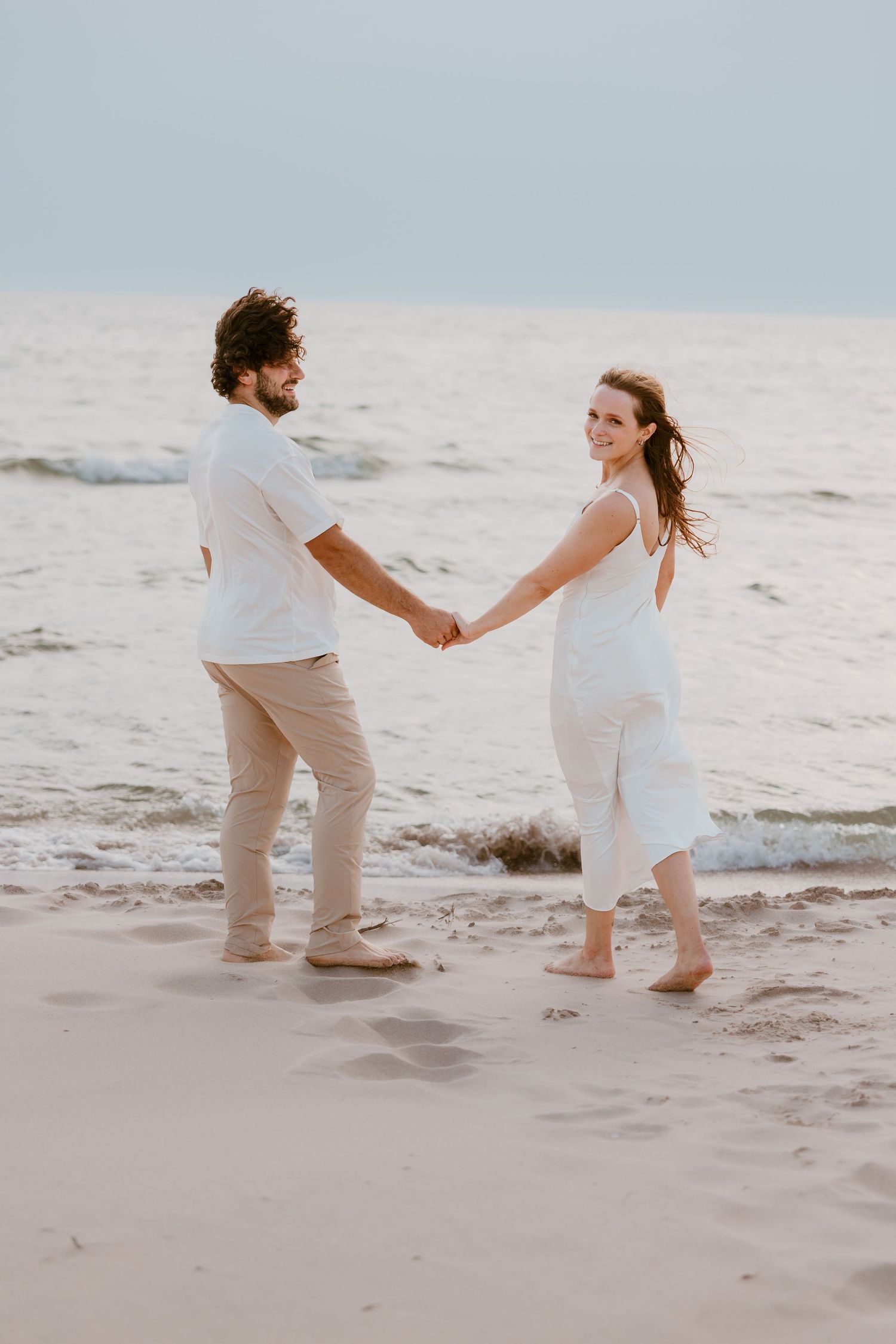 A couple holding hands stands barefoot on a sandy beach at sunset wearing white and beige outfits. Detroit Wedding and Engagement Photo