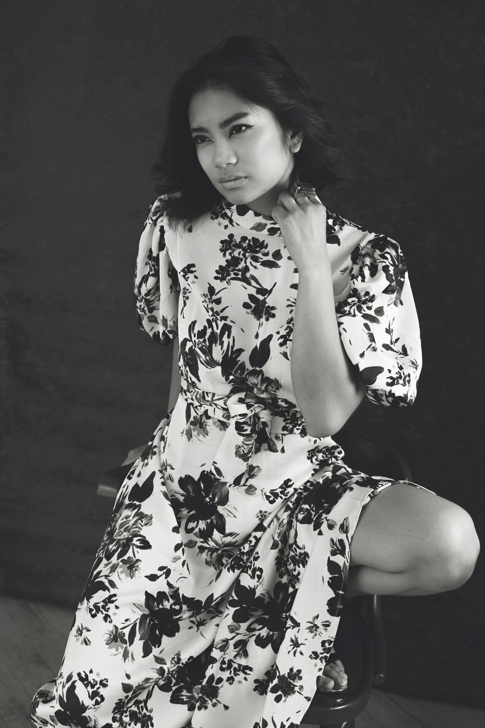 Black and white portrait of someone wearing a floral patterned dress with puff sleeves posing against a dark backdrop.