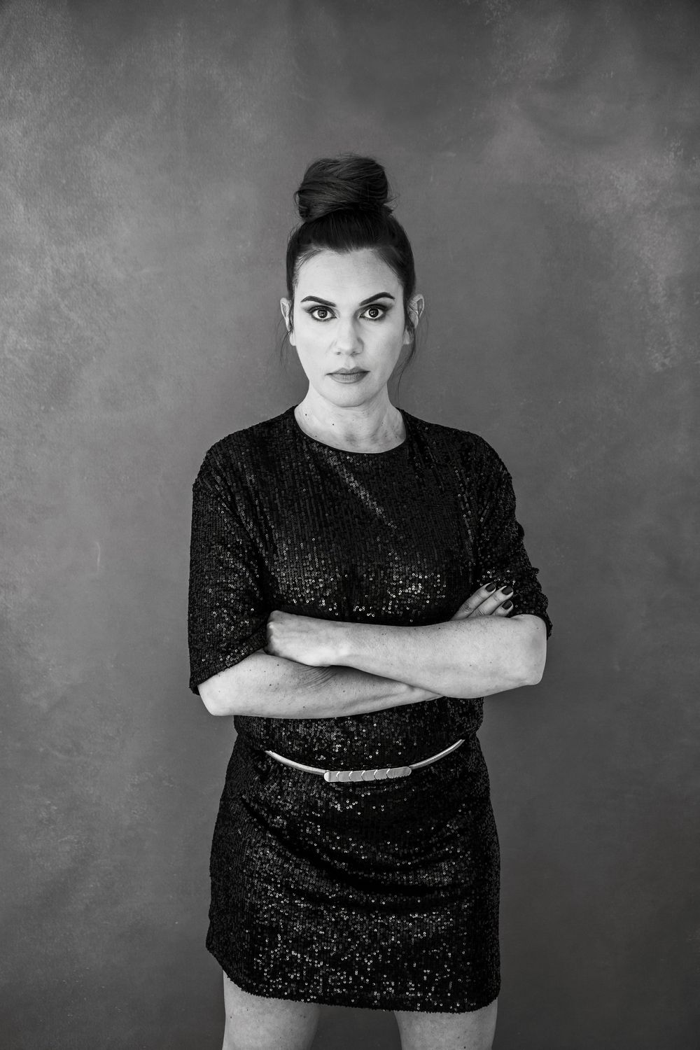 Black and white portrait of elegant woman in sparkling dress with arms crossed and hair in updo against dark backdrop.