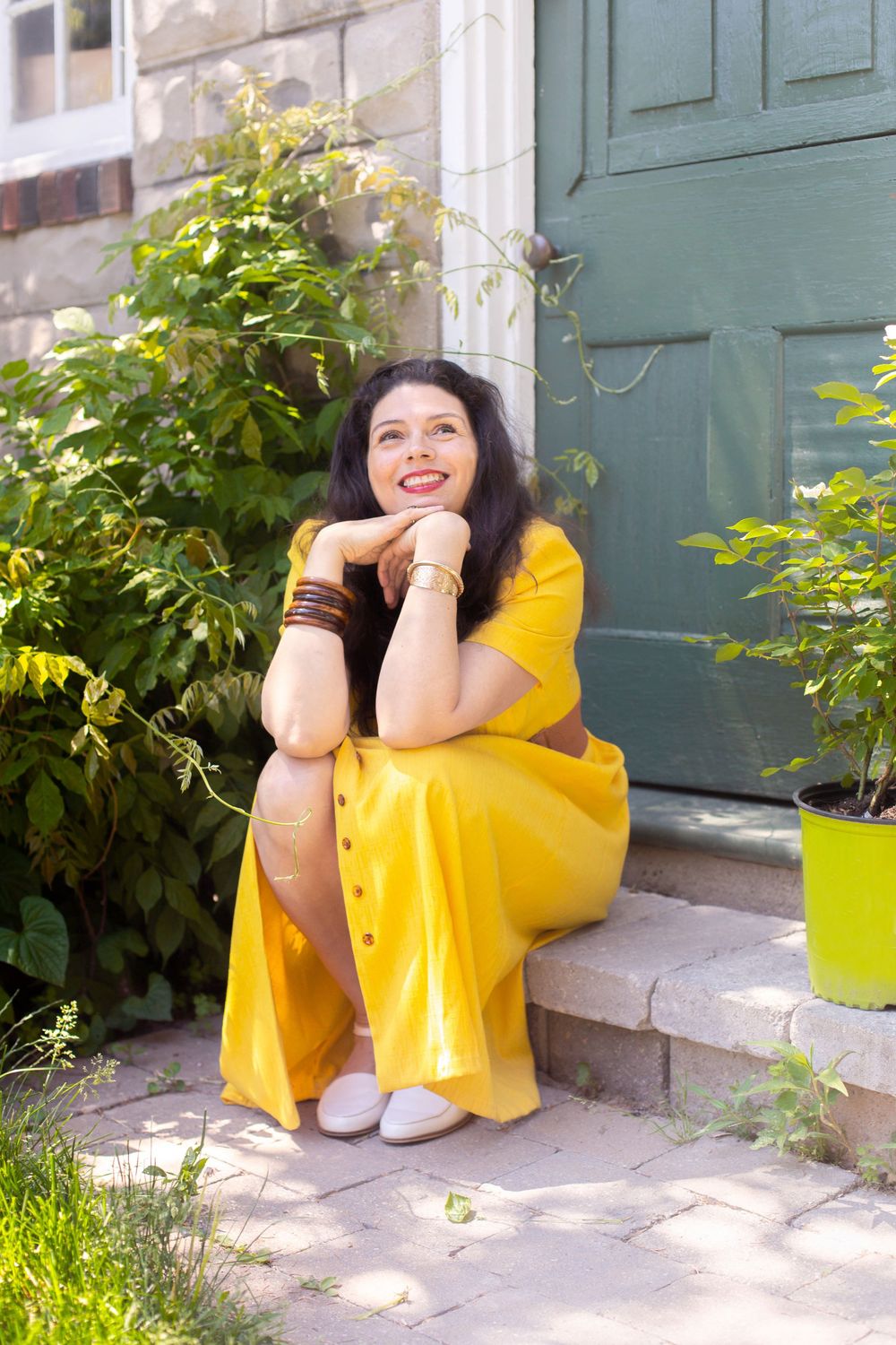 Person wearing a bright yellow dress sits on outdoor steps by a teal door and greenery on a sunny day.