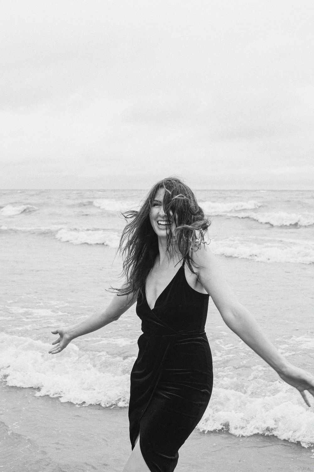A carefree figure in a black dress dances joyfully on a beach with waves crashing behind in black and white photography.