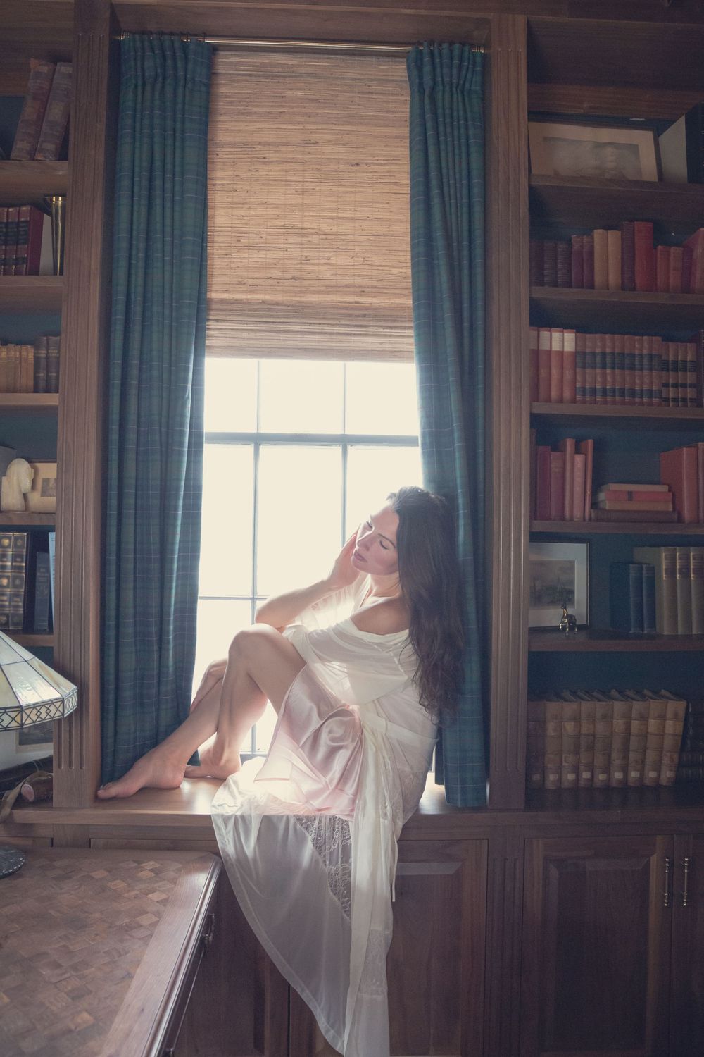 A figure in a white dress poses artistically near bookshelves and a window in a sunlit library setting.