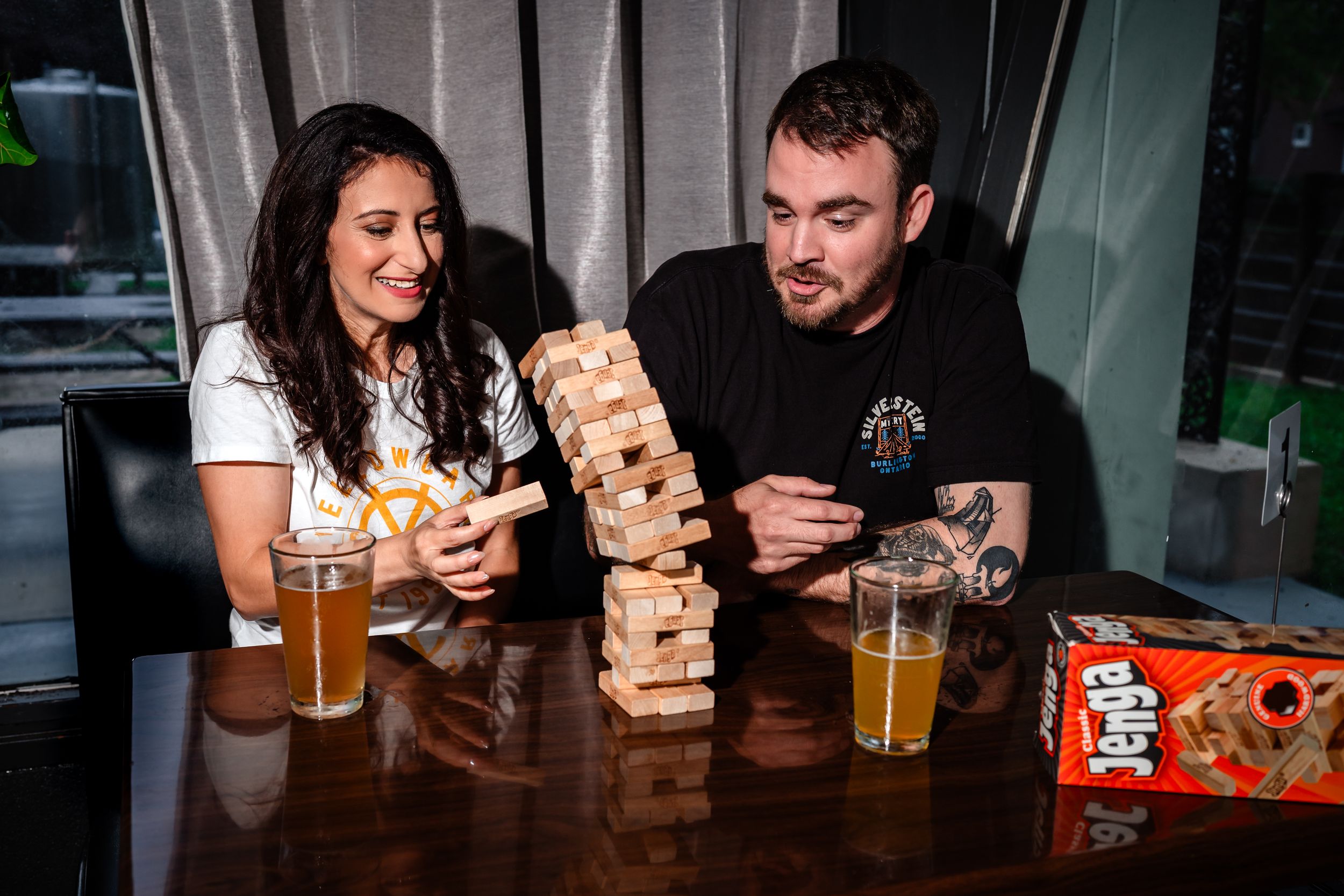 Friends gathered around a table playing Jenga with beers at a casual bar setting.