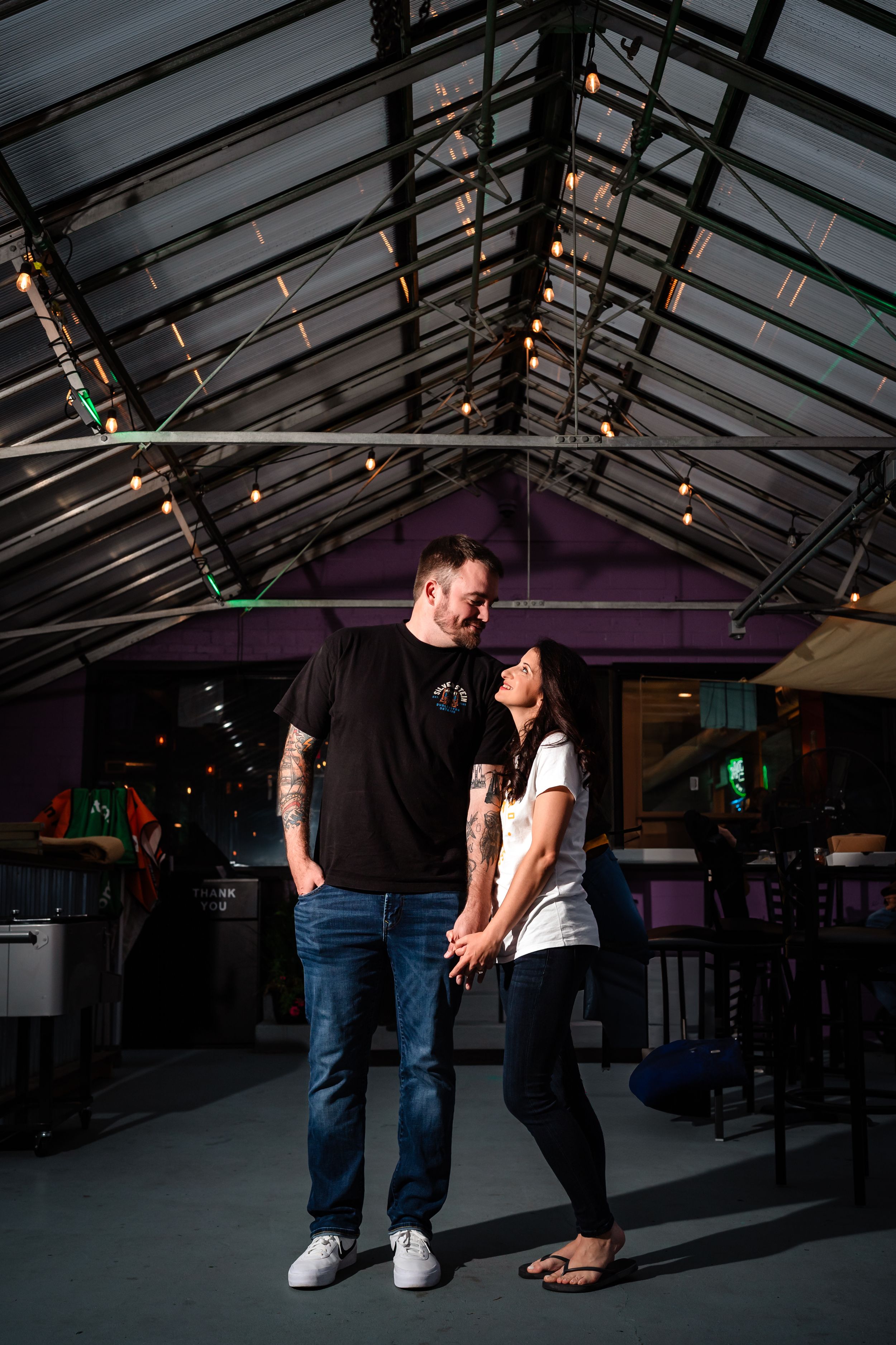 Couple standing in an industrial-style venue with glass ceiling and string lights.