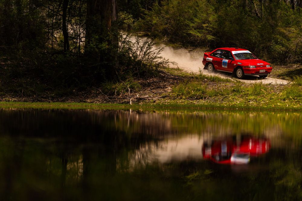 A red sports car drives along a scenic forest road next to a tranquil lake with reflections in the water.