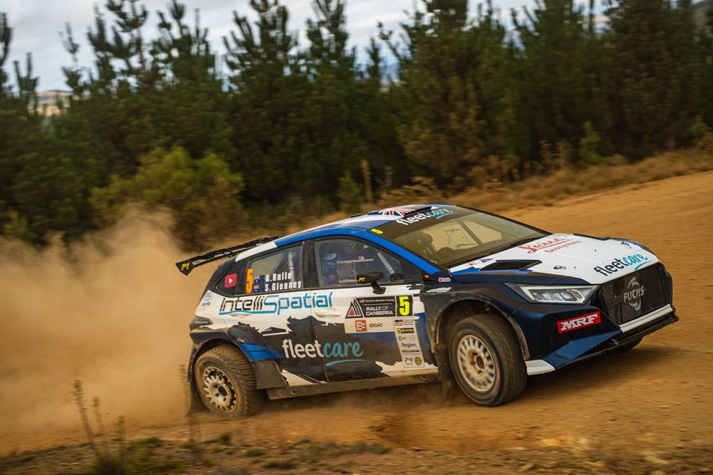 A blue Ford rally car kicks up dust while racing on a dirt track through a forested area.