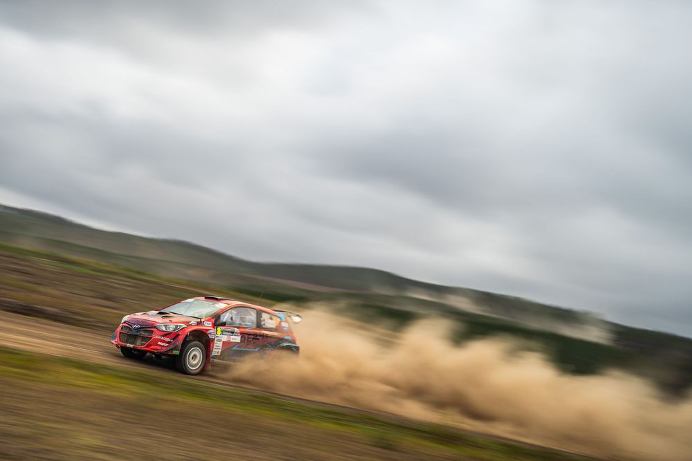 Rally car kicks up dust while racing through green hills under dramatic cloudy skies.
