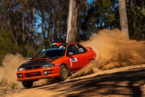 Red BMW rally car kicks up dirt while drifting around a dusty corner during an off-road race.