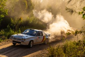 A rally car kicks up dust while racing through a dirt road on a sunny day.