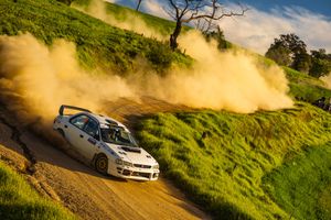 A rally car kicks up dust while racing on a winding dirt track along a green hillside under blue skies.