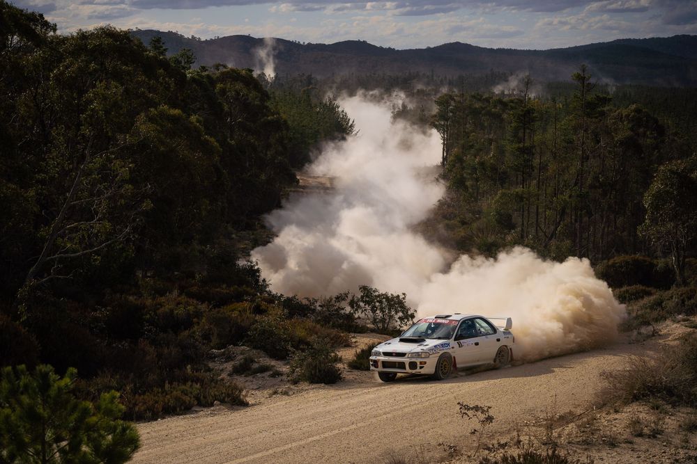 Rally car kicks up dust while racing on a mountain dirt road through forested terrain.