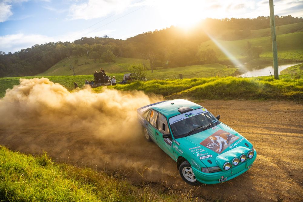 A teal sports car drifts on a dirt track creating dramatic dust clouds during golden hour sunset.