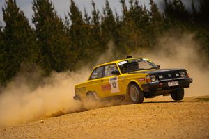 A yellow vintage Volvo rally car kicks up dust while racing on a dirt track with pine trees in the background.