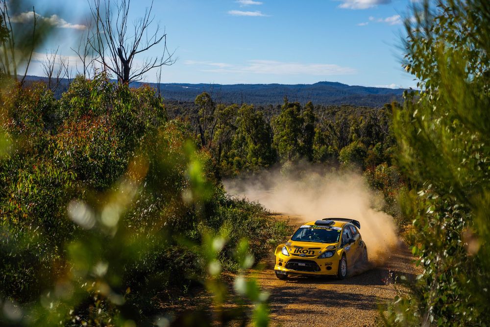 Yellow rally car kicks up dust while racing through a forested mountain course on a sunny day.