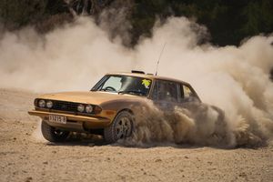 A vintage BMW rally car kicks up dust while drifting through a dirt track during an off-road race.