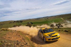 Yellow rally car kicks up dust while racing through a winding dirt track in rugged desert terrain.