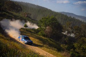 Rally car kicks up dust while racing through a winding mountain course on a sunny day.