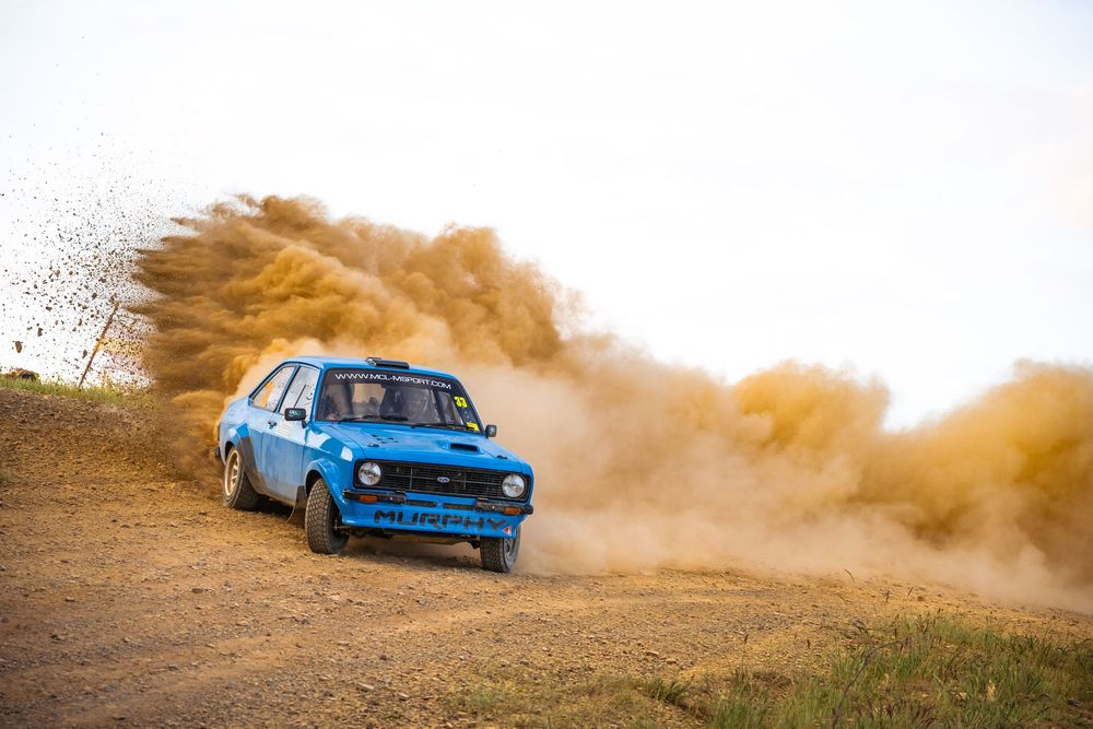 A bright blue rally car kicks up dust while drifting along a dirt road at sunset.