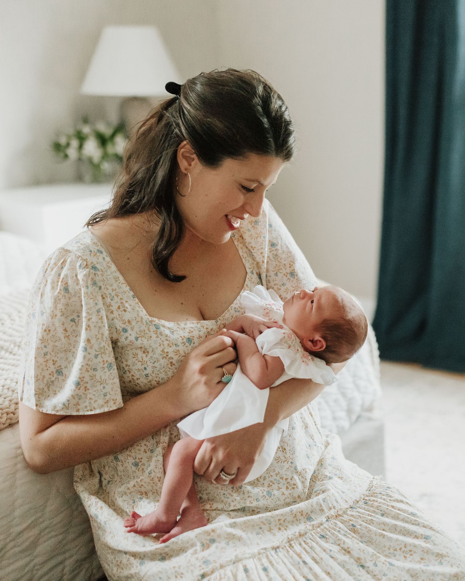 A woman in a floral dress cradles and smiles at her newborn baby in a bright bedroom.