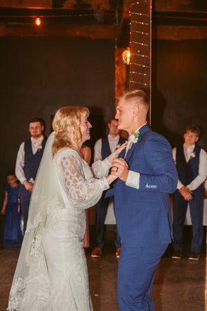 Newlyweds share their first dance at rustic wedding reception under warm lighting and string lights.