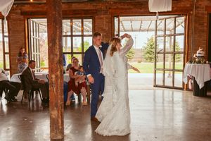 A couple shares their first dance in a rustic barn venue with wooden beams and large glass doors opening to nature.