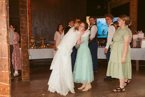 Wedding guests dance together on a rustic wood-floor venue space decorated with string lights and exposed brick walls.