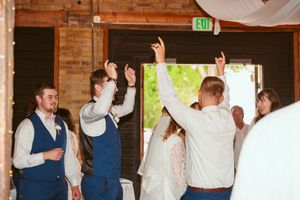 Wedding guests in blue vests and formal attire dancing and celebrating with raised arms in a rustic brick venue.