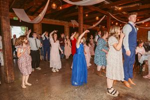 A lively dance party scene at a rustic indoor venue with string lights and people dancing in floral dresses and formal wear.