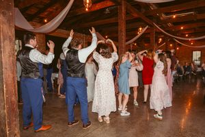 Wedding guests dance and celebrate together on a rustic wooden dance floor with warm lighting and decorative draping overhead.
