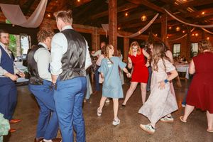 Wedding guests dance together in a rustic wooden barn venue decorated with string lights and white drapes.