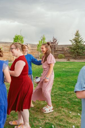 A group of party guests in colorful dresses dance together on a grassy lawn on an overcast day.