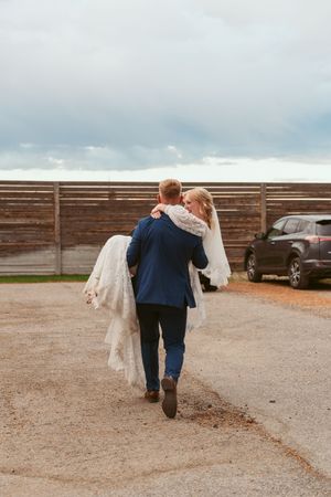 Couple walking away from camera across gravel parking lot beside wooden fence.