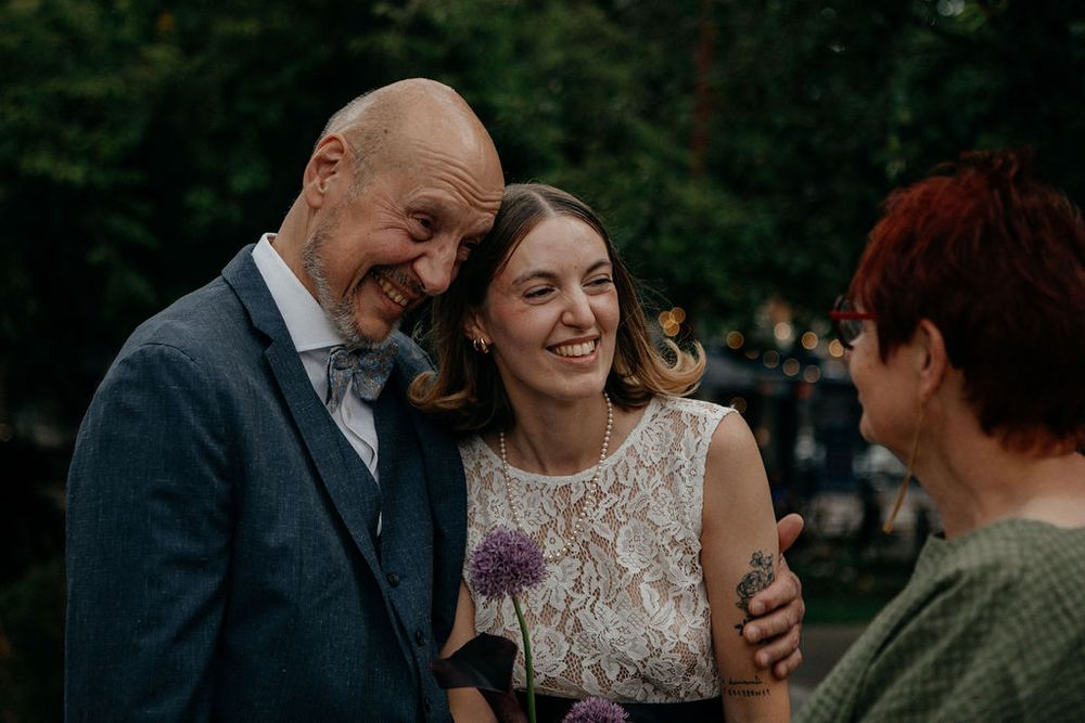 Smiling bride and groom share a joyful moment with a guest at an outdoor wedding reception.