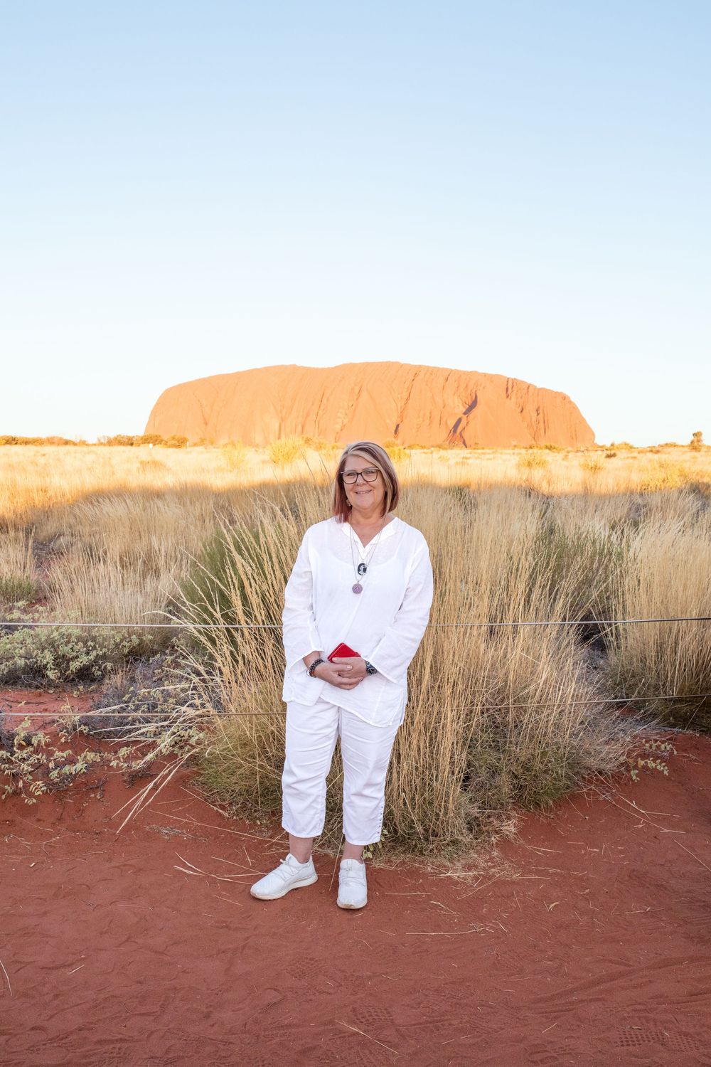 Someone in white clothing poses against the red soil of Uluru with the iconic rock formation visible at sunset.
