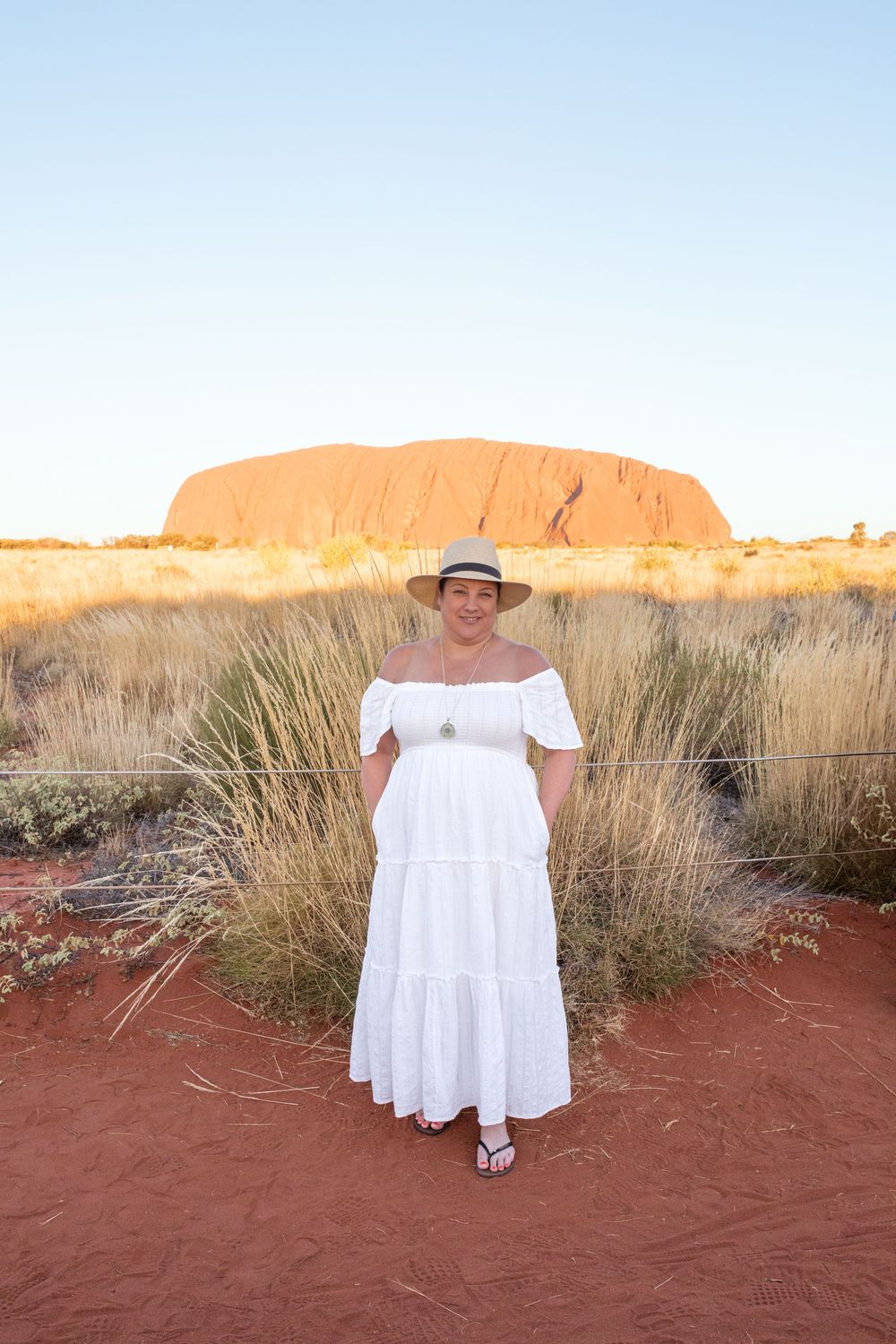 A figure in a white dress stands near desert grasses with Uluru glowing orange in the background.