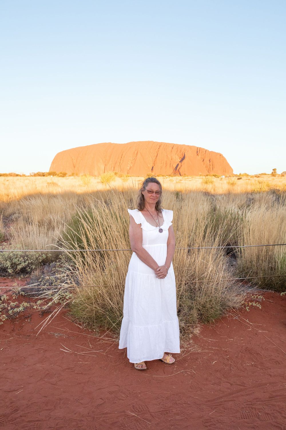 A visitor in a white dress stands against the backdrop of Uluru rock formation in Australia's red desert landscape.