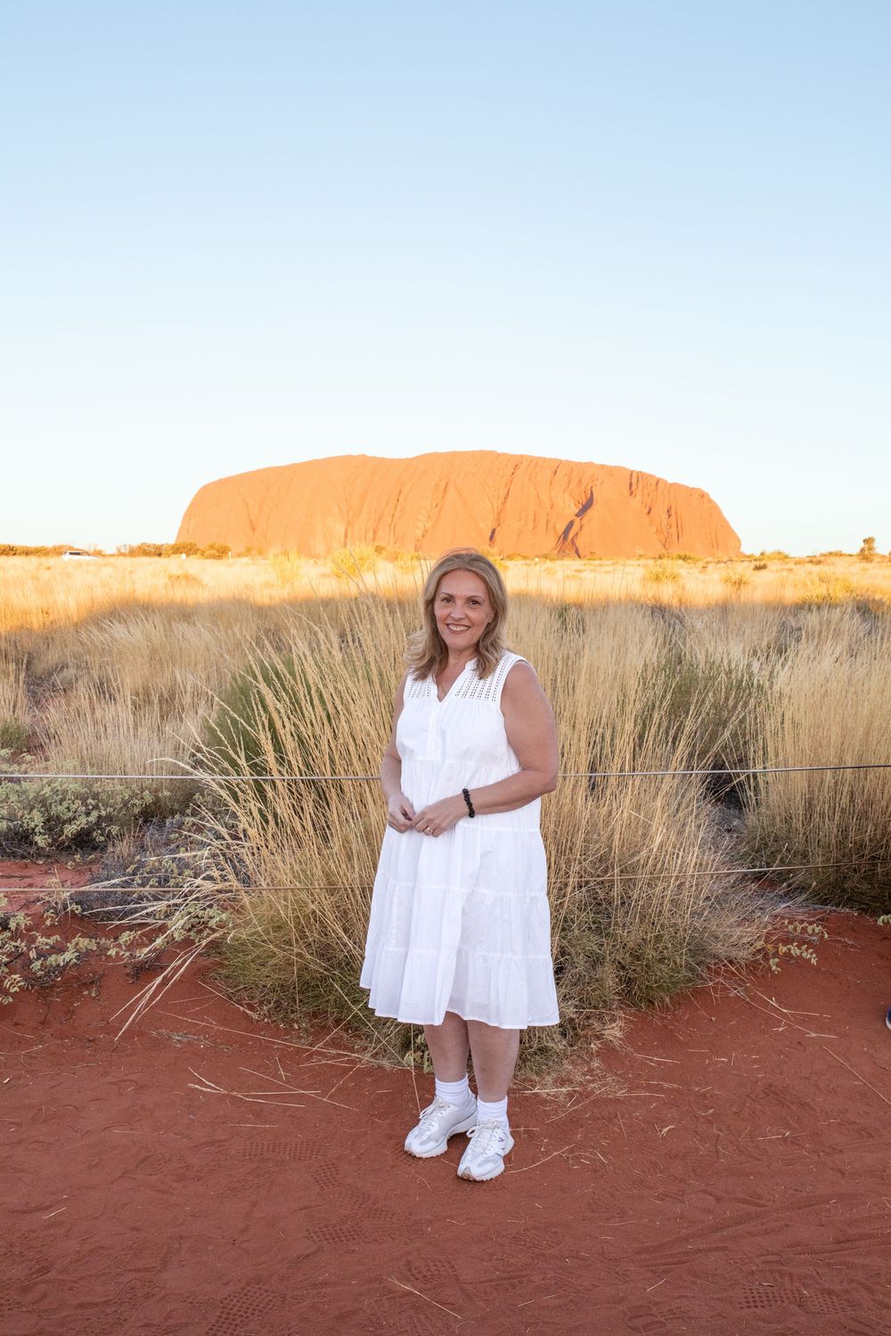 Visitor stands in white dress against iconic Uluru rock formation at sunset in Australia's Red Centre.